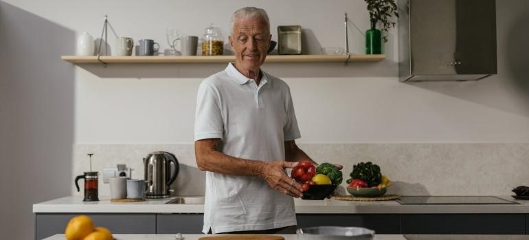 An elderly man is holding a bowl of fruit in the kitchen.